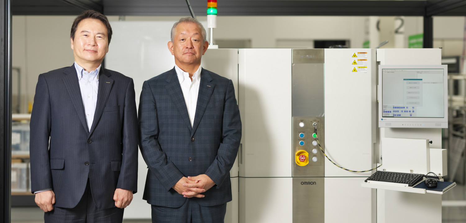 （左）オムロン インダストリアルオートメーションビジネスカンパニー 検査システム事業本部 X線検査システム事業部 開発部 開発2課 課長 益田真之氏　（右）オムロン インダストリアルオートメーションビジネスカンパニー 検査システム事業本部 本部長 渋谷和久氏　※CT型X線自動検査装置「VT-X950」の実機を背景に撮影