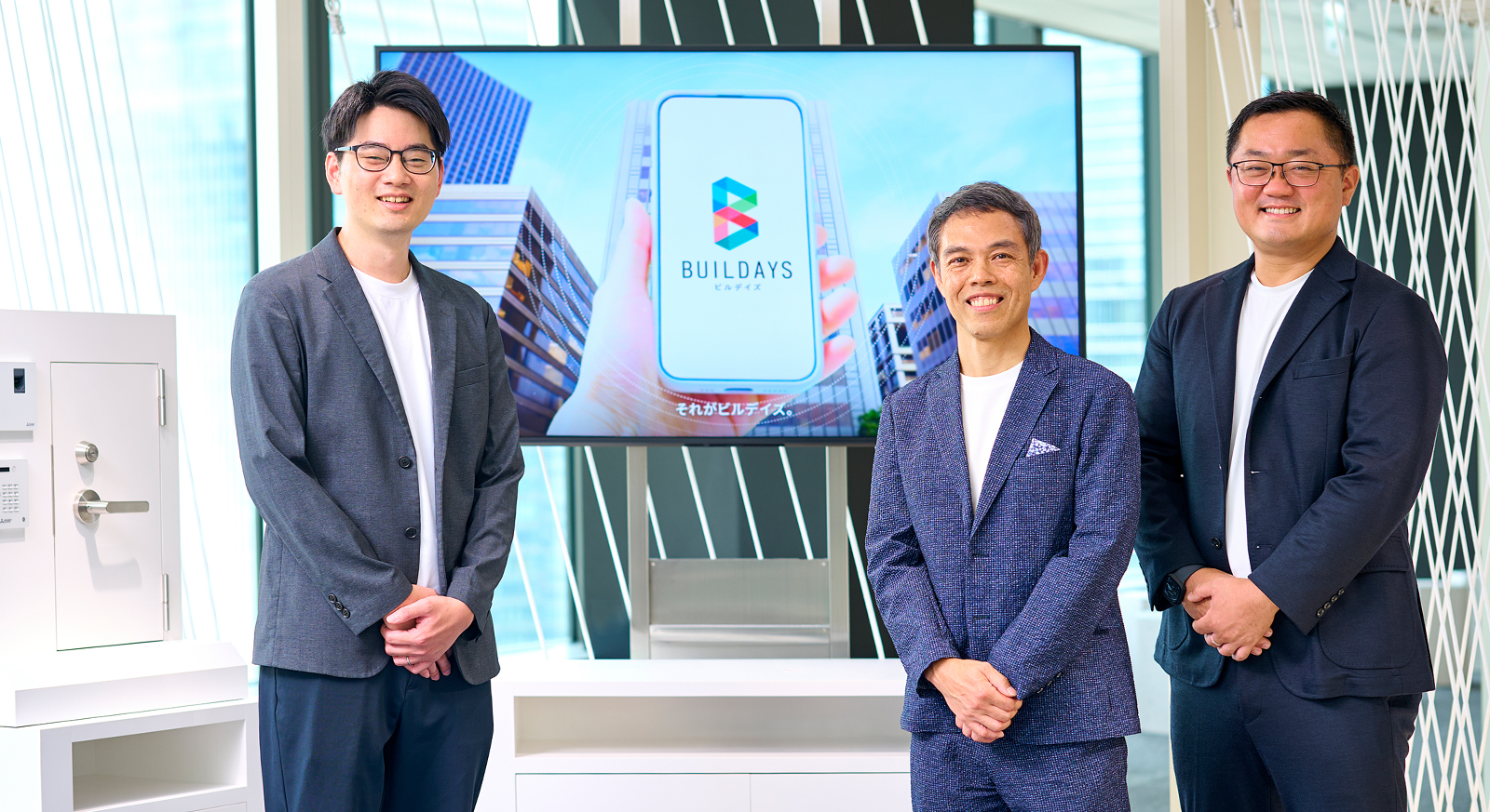 BUILDAYSの画面を背景に並ぶ平井友樹氏、塩原義浩氏、甲斐正太郎氏